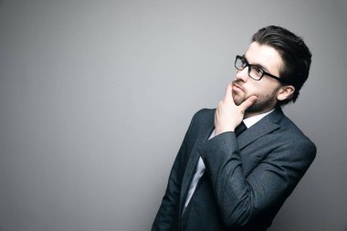 Takım elbiseli ve gözlüklü bir adam gri arka planda çenesine dokunur ve düşünür.