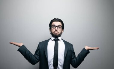Takım elbiseli ve gözlüklü bir adam gri arka planda şaşırarak ellerini açtı.