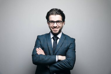 Takım elbiseli ve gözlüklü bir adam gri bir arka planda kollarını kavuşturmuş duruyor.