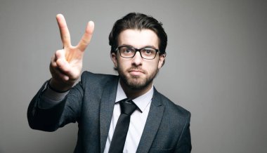Takım elbiseli ve gözlüklü adam gri arka planda barış işareti gösteriyor.