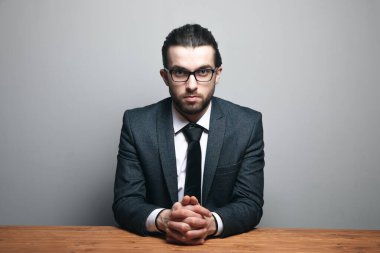 Takım elbiseli ve gözlüklü bir adam gri arka planda oturuyor.