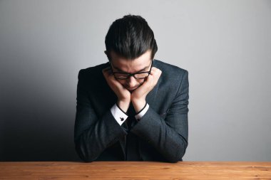 Takım elbiseli ve gözlüklü bir adam elleri çenesinin altında gri arka planda oturuyor.