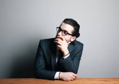 Takım elbiseli ve gözlüklü bir adam oturur ve gri bir arka plana bakar.