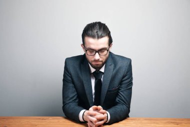 Takım elbiseli ve gözlüklü bir adam gri arka planda kapalı gözlerle oturuyor.