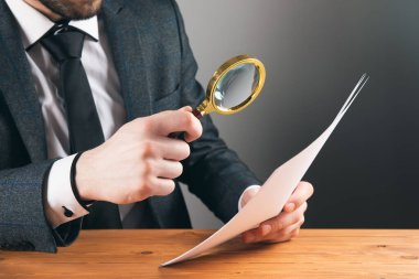 Büyüteci olan takım elbiseli bir adam gri arka plandaki bir kağıda bakıyor.