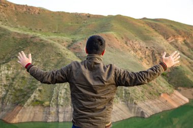Bir adam açık ellerle gölün kıyısındaki bir uçurumda duruyor.