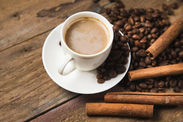 a cup of coffee, coffee seeds, cinnamon and a book on a wooden table