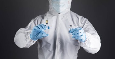 Koruyucu üniformalı doktor gri arka planda bir şırınga ve ampul tutuyor.