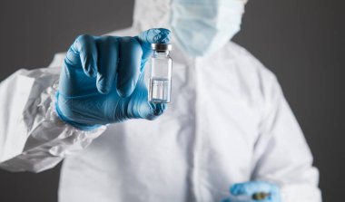 Koruyucu üniformalı doktor gri arka planda bir şırınga ve ampul tutuyor.