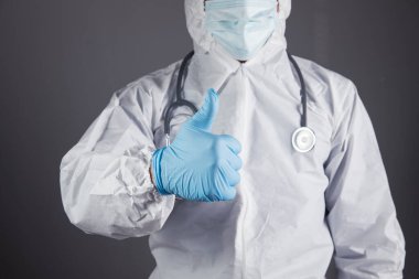 Beyaz koruyucu üniformalı doktor gri arka planda baş parmağını gösteriyor.