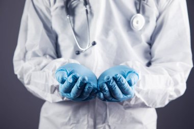 Koruyucu üniformalı doktor avuç içi açık gösteriyor. Gri bir arka planda duran eller.