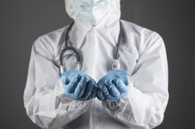 Koruyucu üniformalı doktor avuç içi açık gösteriyor. Gri bir arka planda duran eller.