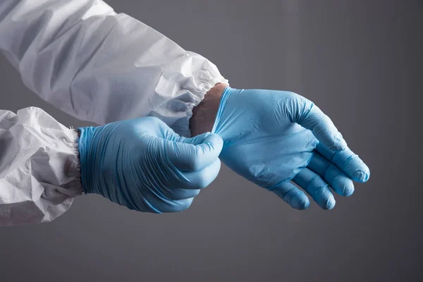 Beyaz koruyucu üniformalı bir doktor gri arka planda bir eldiven giyer.