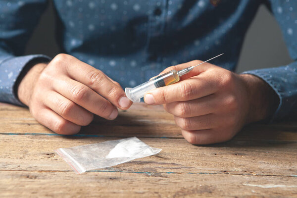 the addict is holding a syringe with liquid and a bag of powder on the table. addict concept