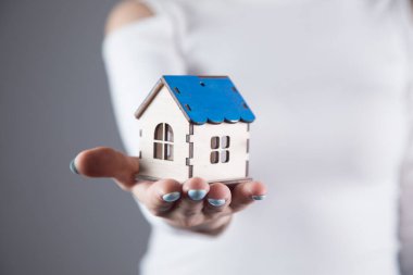 Genç bir kadın, gri bir arka planda elinde bir ev tutuyor.