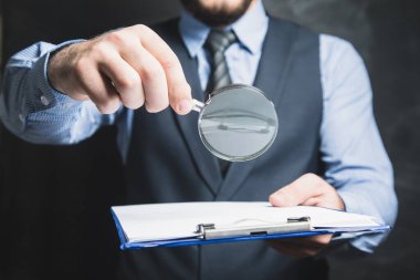 Takım elbiseli bir adam gri arka planda büyüteçle belgelere bakıyor.