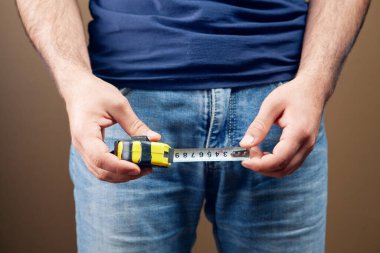 a man holds a centimeter in his hands and measures a penis on a brown background