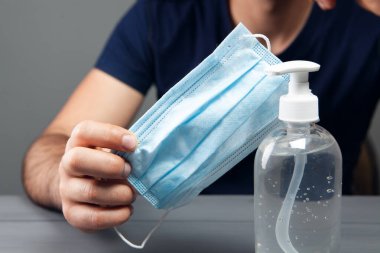 Antiseptiğin önünde otururken elinde maske tutan adam gri arka planda.