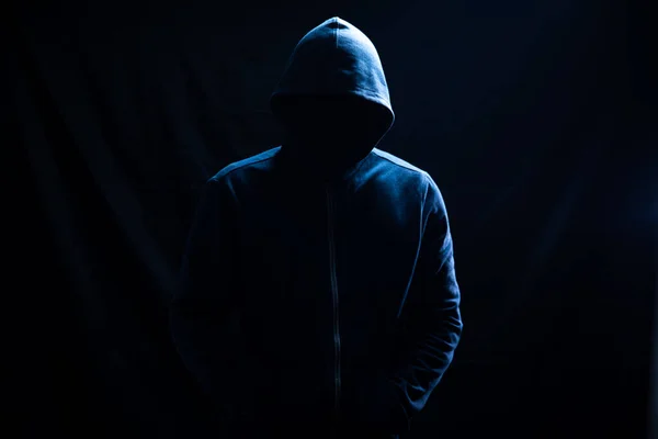 Kapüşonlu bir adam siyah arka planda duruyor.
