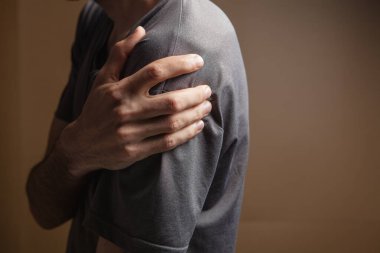 Genç bir adamın, kahverengi arka planda omuz ağrısı vardır.