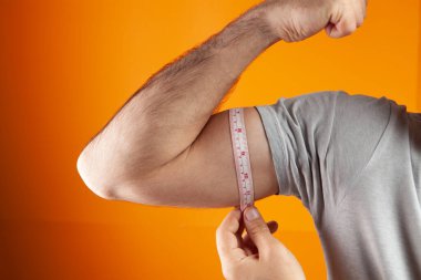 Bir adam kolunun kalınlığını turuncu arka planda bir metreyle ölçer.