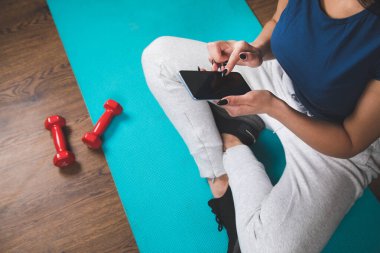 Bir kadın egzersiz minderinde otururken telefonu tutuyor.