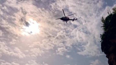Bir kurtarma helikopteri kayalık sahil boyunca mavi denizin üzerinde alçaktan uçuyor.