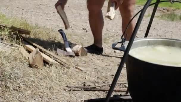 Egy férfi fát vág egy fazék hátterében a tűzön. — Stock videók