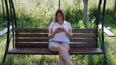 Bir kadın akıllı telefona bakar, bahçede salıncakta sallanır..