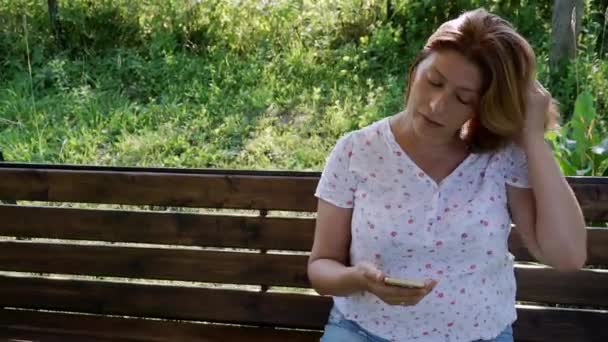 Una mujer mira en un teléfono inteligente, balanceándose en un columpio en el jardín. — Vídeos de Stock