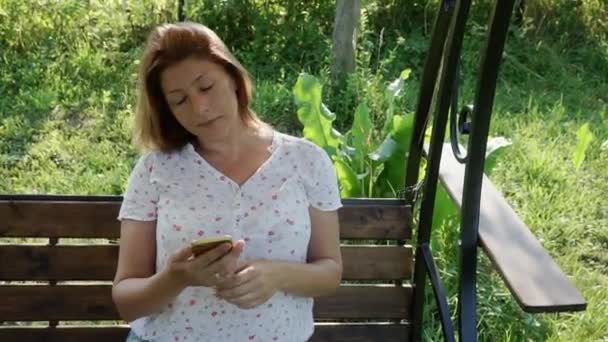 Kobieta patrzy na smartfona, huśtając się na huśtawce w ogrodzie. — Wideo stockowe