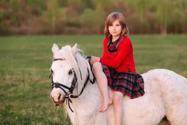 Giovane ragazza in abito rosso seduta su un pony bianco — Foto Stock