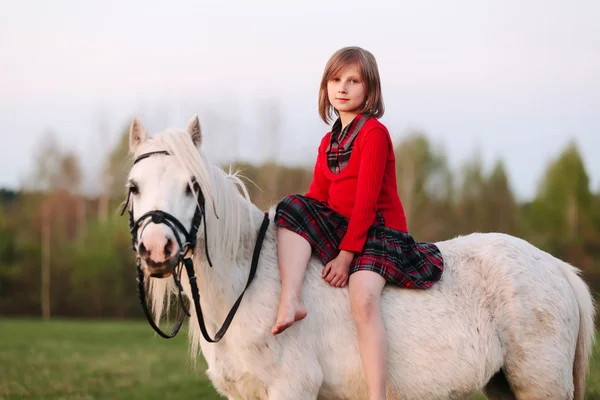 Menina criança está sentada em um pequeno cavalo branco olhando para a câmera — Fotografia de Stock