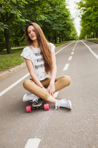 Attractive young woman sitting cross-legged on a longboard on the road in the park. Skateboarding. Outdoors — Stock Photo, Image