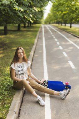 Genç kız öğrenci bir gömlek ve pantolon longboard yanındaki parkta bir halı üzerinde oturuyor. Kaykay. Açık havada