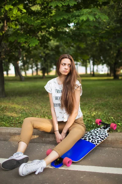 Den unge pige med et trist ansigt sidder i parken med en longboard. udendørs, livsstil - Stock-foto