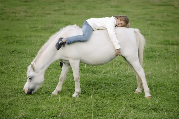 Niña pequeña en un suéter blanco y vaqueros acostados al revés en la parte posterior de un caballo blanco. Retrato de estilo de vida —  Fotos de Stock