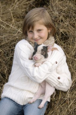 Young girl sitting on hay, smiling and holding a pig in his smiles. Lifestyle portrait clipart
