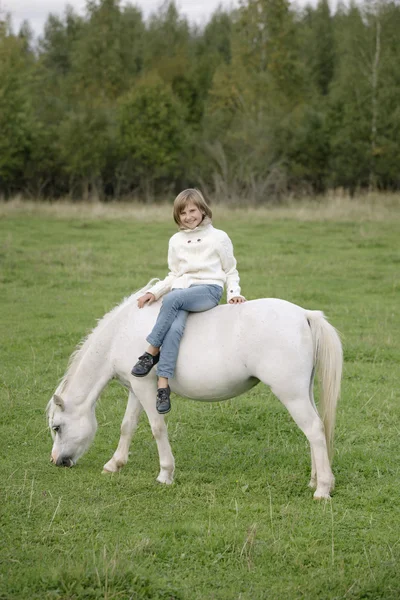 Jeune fille en pull blanc et jean assis les jambes croisées sur un cheval blanc. Portrait de style de vie — Photo