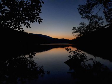 Hırvatistan 'da akşam gökyüzüne karşı ağaç ve dağ siluetiyle gölde alacakaranlık