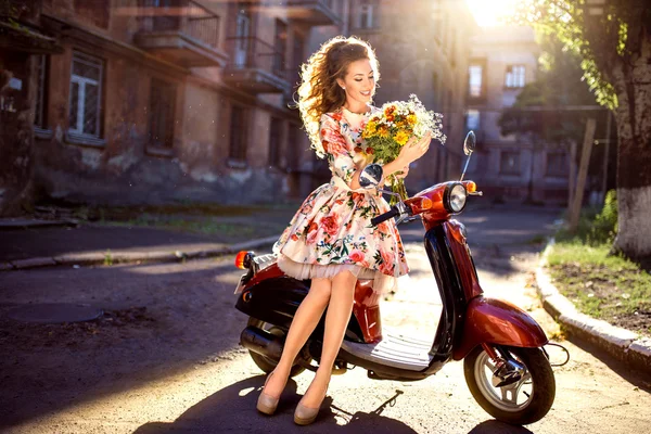 Beautiful middle-aged woman with flowers and curly hair sits on a scooter in a dress. In the rays of the sun — Stock Photo, Image