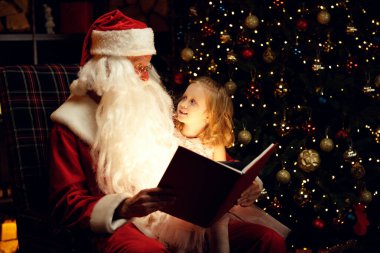 Noel Baba ve mutlu küçük bir kız sihirli bir kitap izliyor Noel Baba 'nın kucağında oturan bir çocuk