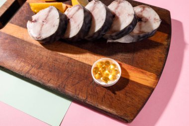 Taze doğranmış uskumru, parlak renkli arka planda morina yağı kapsülleriyle birlikte mutfak tahtasının üzerinde yatıyor.