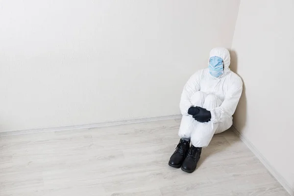 A doomed man in a protective suit and many medical masks on his face sits in the corner of the room. He hides his entire face to protect himself from the coronavirus.