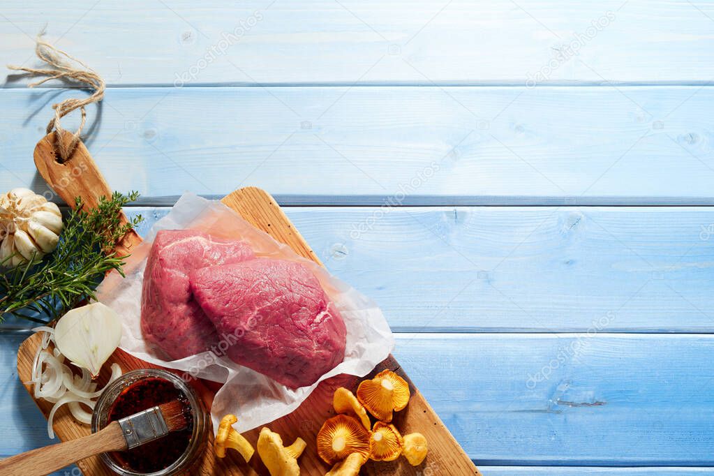 Overhead view of raw beef joint with chanterelle mushrooms and ingredients, light blue wood background with copy space