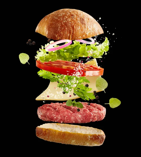 Floating fresh ingredients for a beef burger over a black background with fresh salad greens, bun, condiments , slice of cheese and raw meat patty suspended midair in layers