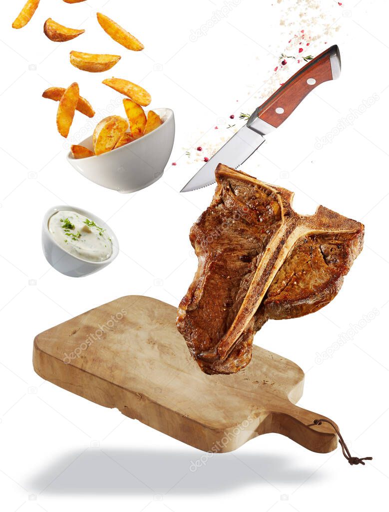 Grilled T-bone steak floating ingredients concept with a chopping board, healthy lean steak, French fries and tzatziki in bowls, spices and a sharp knife isolated on white