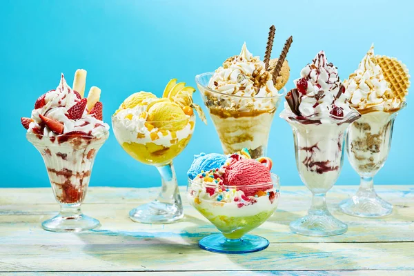 Five different flavor ice cream sundaes on light wooden table with blue background