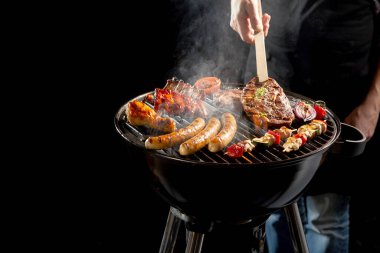 Man grilling an assortment of meat and kebabs on a portable barbecue lifting a t-bone steak with a pair of tongs in a close up view of his hand clipart