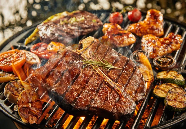 T-bone steak grilling over a portable barbecue outdoors in summer with garlic, tomato, rump steak, chicken wings and baby narrow